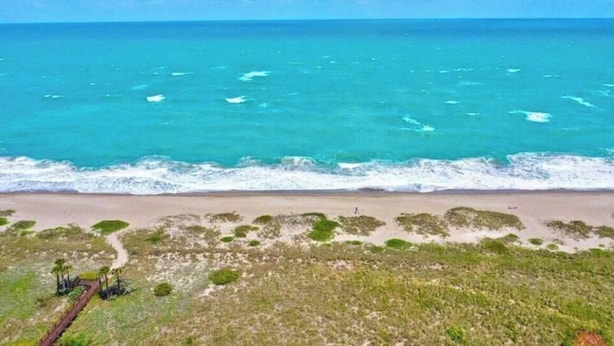 A beach with clear turquoise water, a sandy shore, and sparse vegetation. - Luxurious Beach Resort Atmosphere Getaway (Fort pierce)