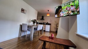 Ein modernes Wohnzimmer mit einem Holzesstisch, einem an der Wand montierten Fernseher und einer Küche im Hintergrund.