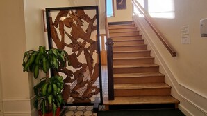 Eine Treppe mit einer Holzhandläufe und einer schwarzen Matte am Fuß. Eine dekorative Holzvertäfelung befindet sich links, daneben ein Blumentopf.