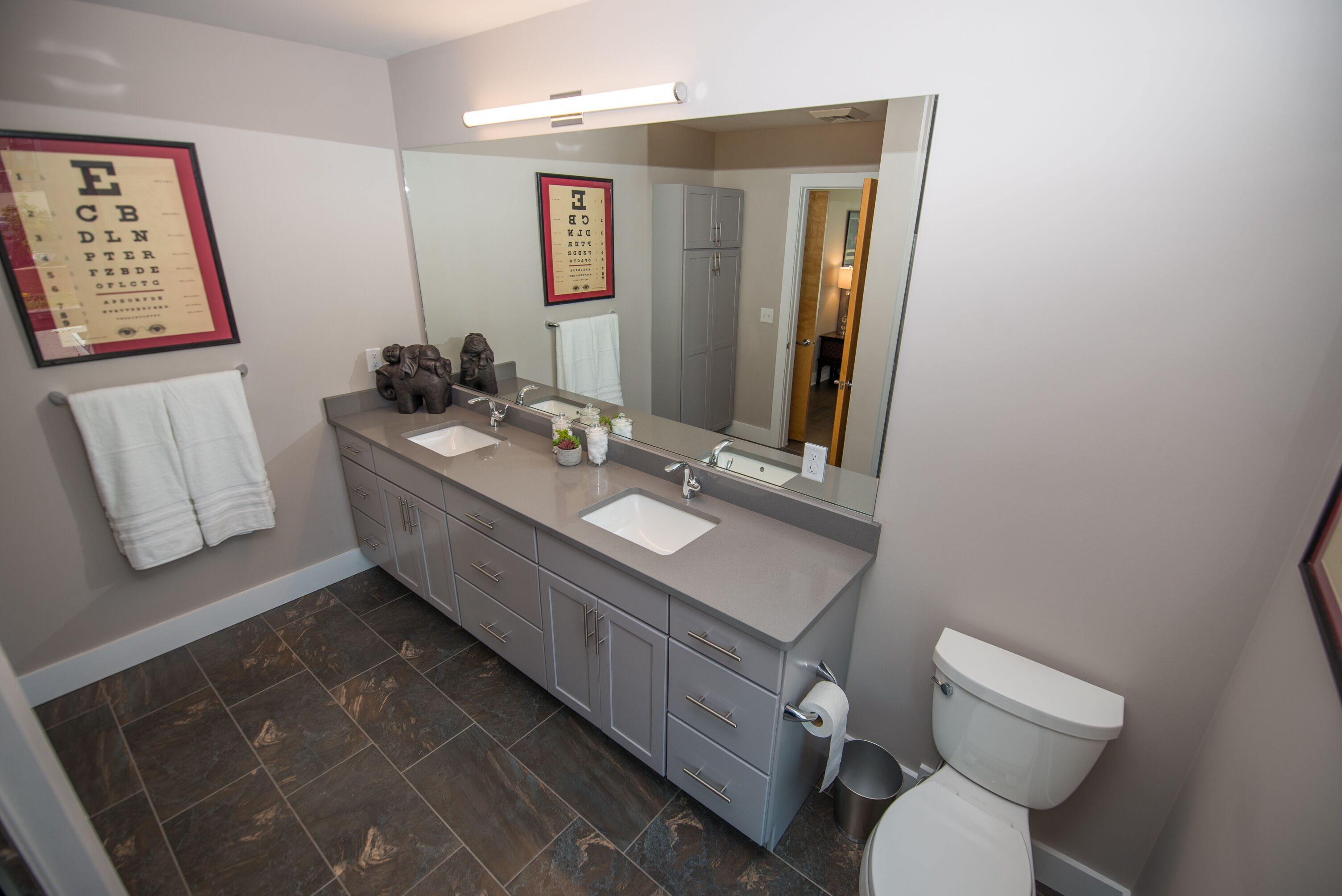Een moderne badkamer met twee wastafels, een grote spiegel en een toilet. Op de muur hangen gekaderde visuele prikkeldagen.