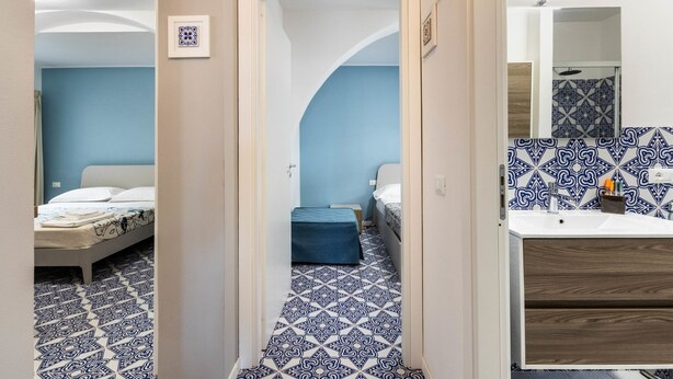 A hotel room with a blue and white patterned floor, a bed with a white headboard, a small desk, and a bathroom with a mirror and patterned tiles.