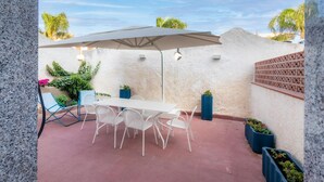 An outdoor patio with a table and chairs under a large umbrella, surrounded by potted plants and a textured wall.