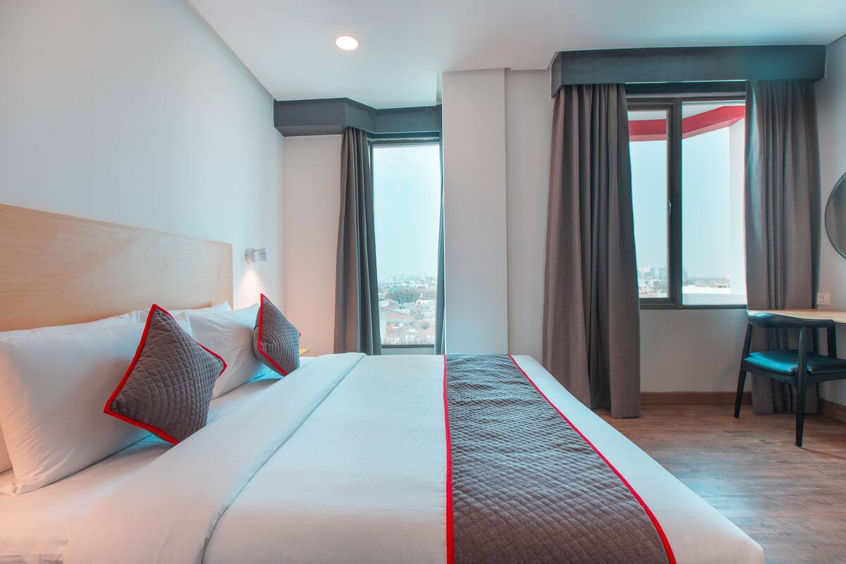 A hotel room with a large bed, a desk, and a view of the city through the window.