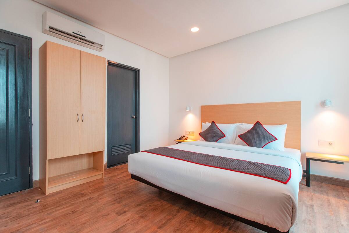 A hotel room with a large bed, wooden wardrobe, bedside tables, and an air conditioning unit.