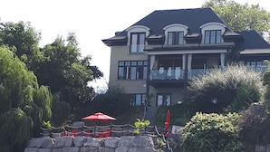 A large, multi-story house with a stone retaining wall in front, featuring a balcony with red umbrellas.