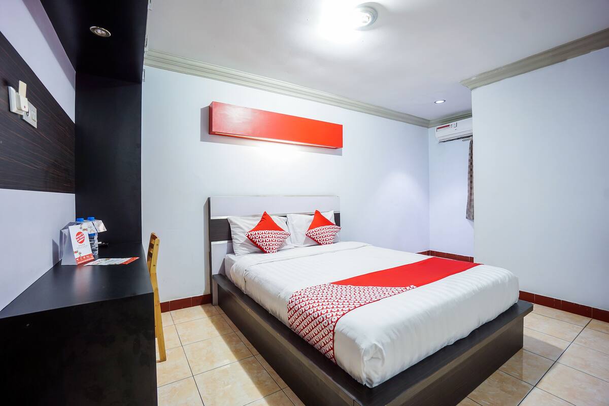 Habitación de hotel con cama, mesita de noche, colcha roja y blanca, y aire acondicionado de pared.