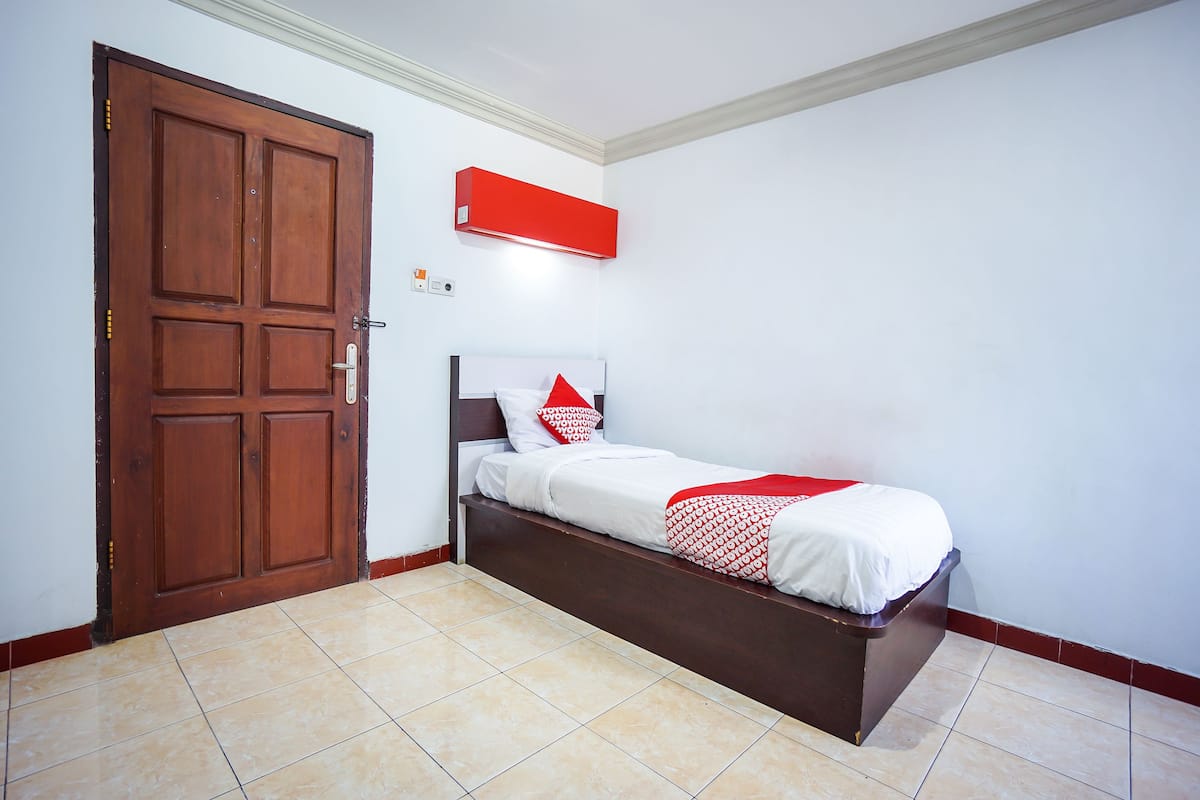 Un dormitorio con puerta de madera, cama con ropa de cama blanca y detalles rojos, y un equipo de aire acondicionado rojo en la pared.