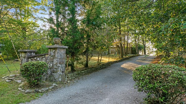 Un sendero de grava serpentea por una zona boscosa con muros de piedra y una urna decorativa sobre un pedestal.