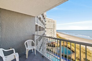 A balcony with two white plastic chairs, a view of a beach, and a pool.