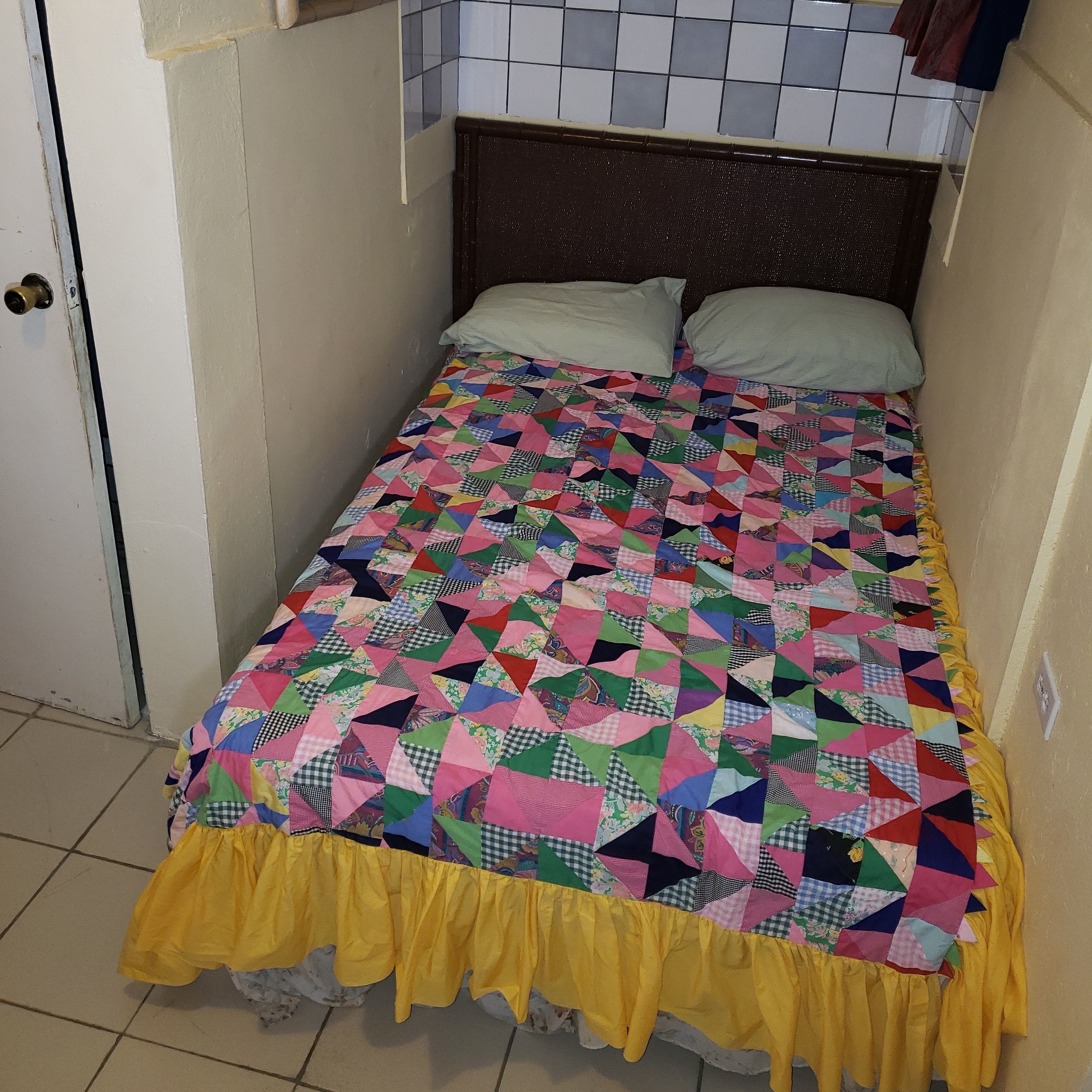 Una habitación pequeña con cama individual, colcha de retazos de colores y ribete amarillo.