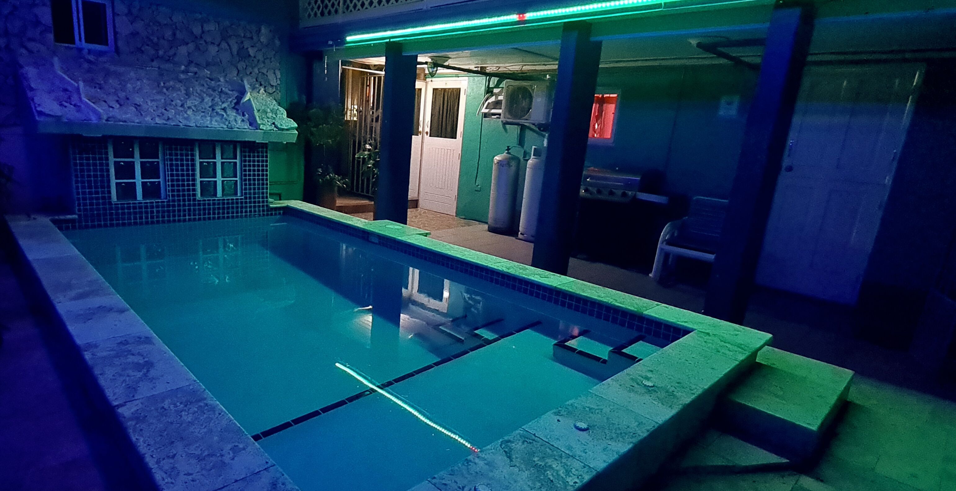 Une piscine avec une terrasse carrelée, un mur en pierre et un éclairage néon vert.