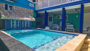 A swimming pool with a stone wall and blue tiles on one side, a covered seating area with chairs, and a green building with a white door. - A1 Apartments Aruba (Oranjestad)