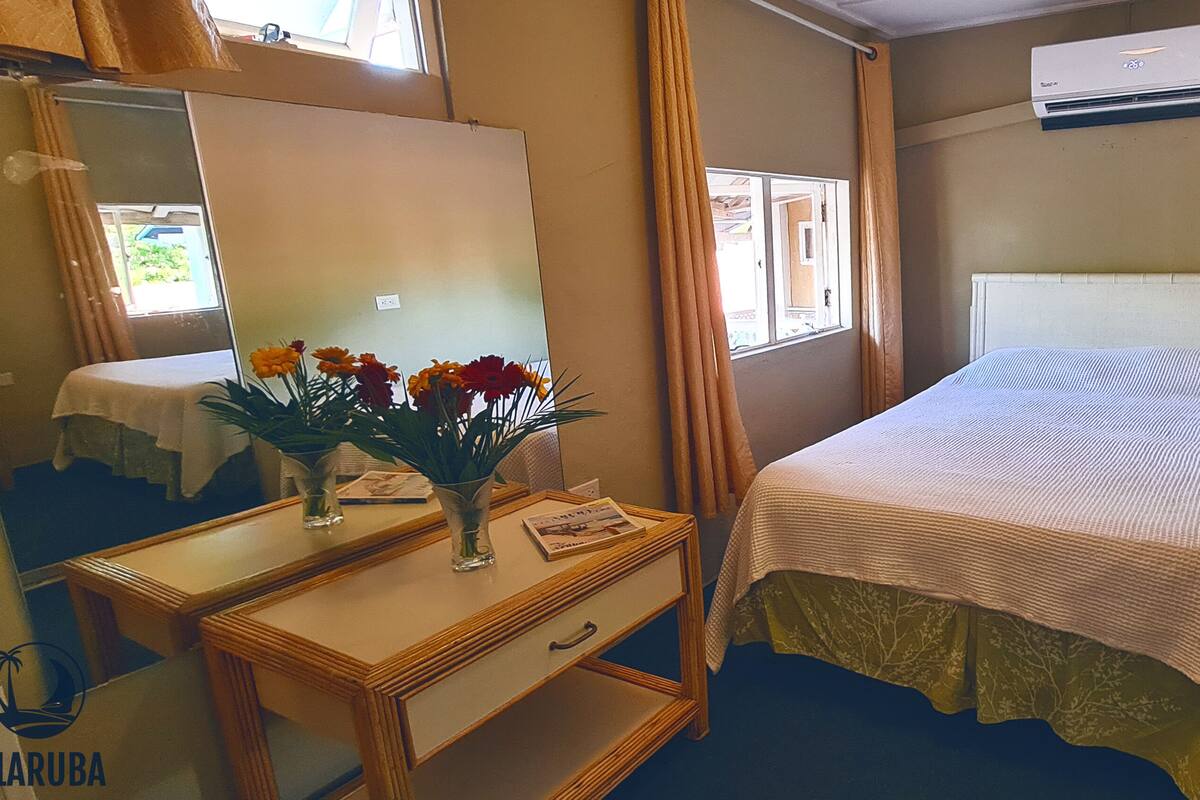 Quarto de hotel com uma cama, uma mesinha de cabeceira com um vaso de flores, um espelho e um aparelho de ar condicionado.