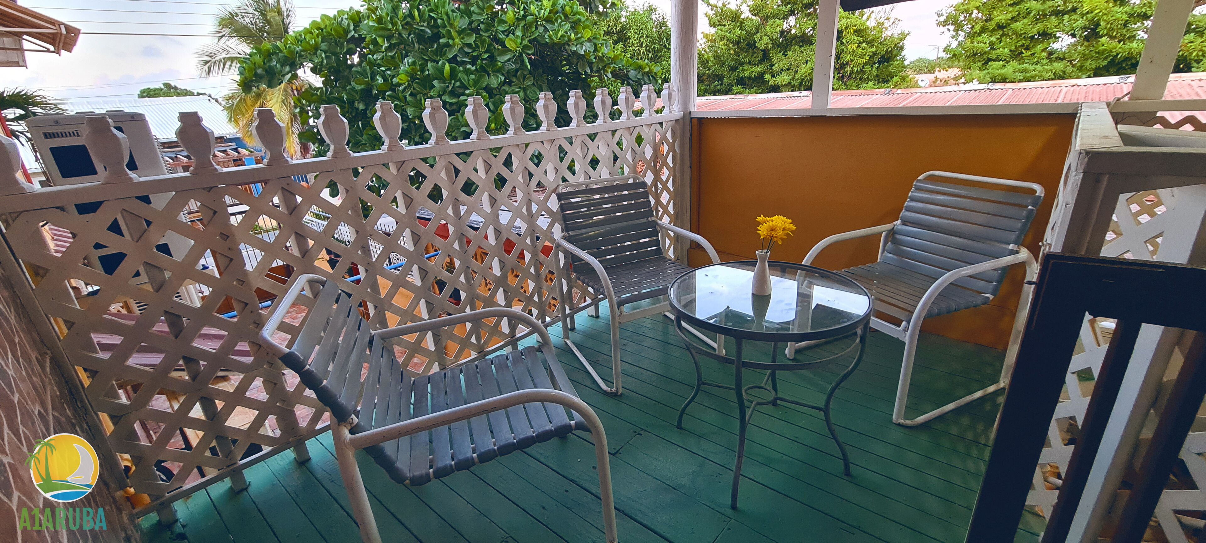 Un balcon avec une balustrade blanche en treillis, une table en verre ornée d’un vase de fleurs jaunes et deux chaises en métal.