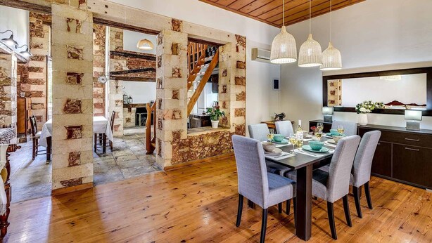A dining area with a wooden table set for four, surrounded by grey chairs. A staircase leads to an upper level, and a kitchen area is visible in the background. - Casa Veneziana Chania, Casa Veneziana (Chania)