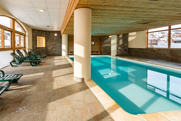 An indoor swimming pool with stone walls and wooden beams, surrounded by lounge chairs and large windows.