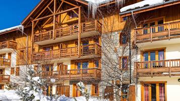 Un chalet en bois doté de balcons, entouré d’un paysage enneigé, et surplombé d’un ciel bleu azur.