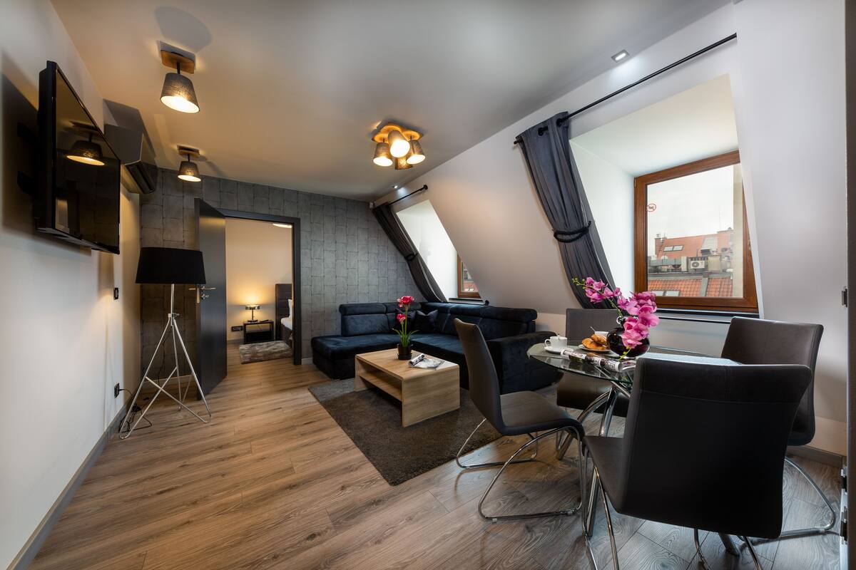 A modern living room with a sofa, coffee table, and dining area. There is a large window with curtains, a flat-screen TV mounted on the wall, and a tripod lamp.