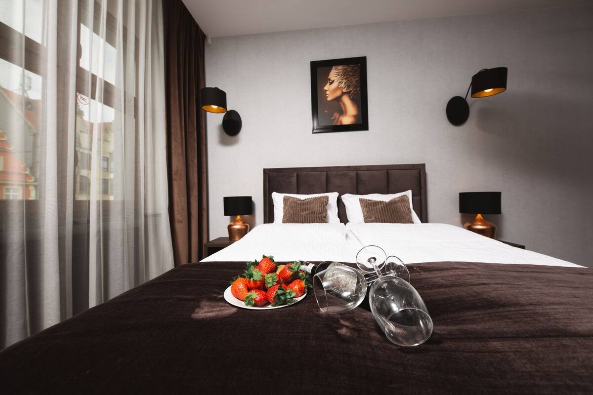 A neatly made bed with a dark brown headboard, two bedside lamps, a framed portrait on the wall, and a plate of strawberries on the bed.