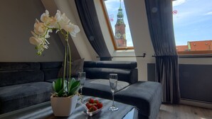 A modern room with a dark gray sofa, a small table with a potted orchid, a bowl of strawberries, and two champagne flutes. A window reveals a clock tower outside. - Aparthotel New Lux (Wroclaw)