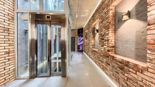 A modern hallway with a brick wall, a glass elevator, and a light fixture. - Aparthotel New Lux (Wroclaw)
