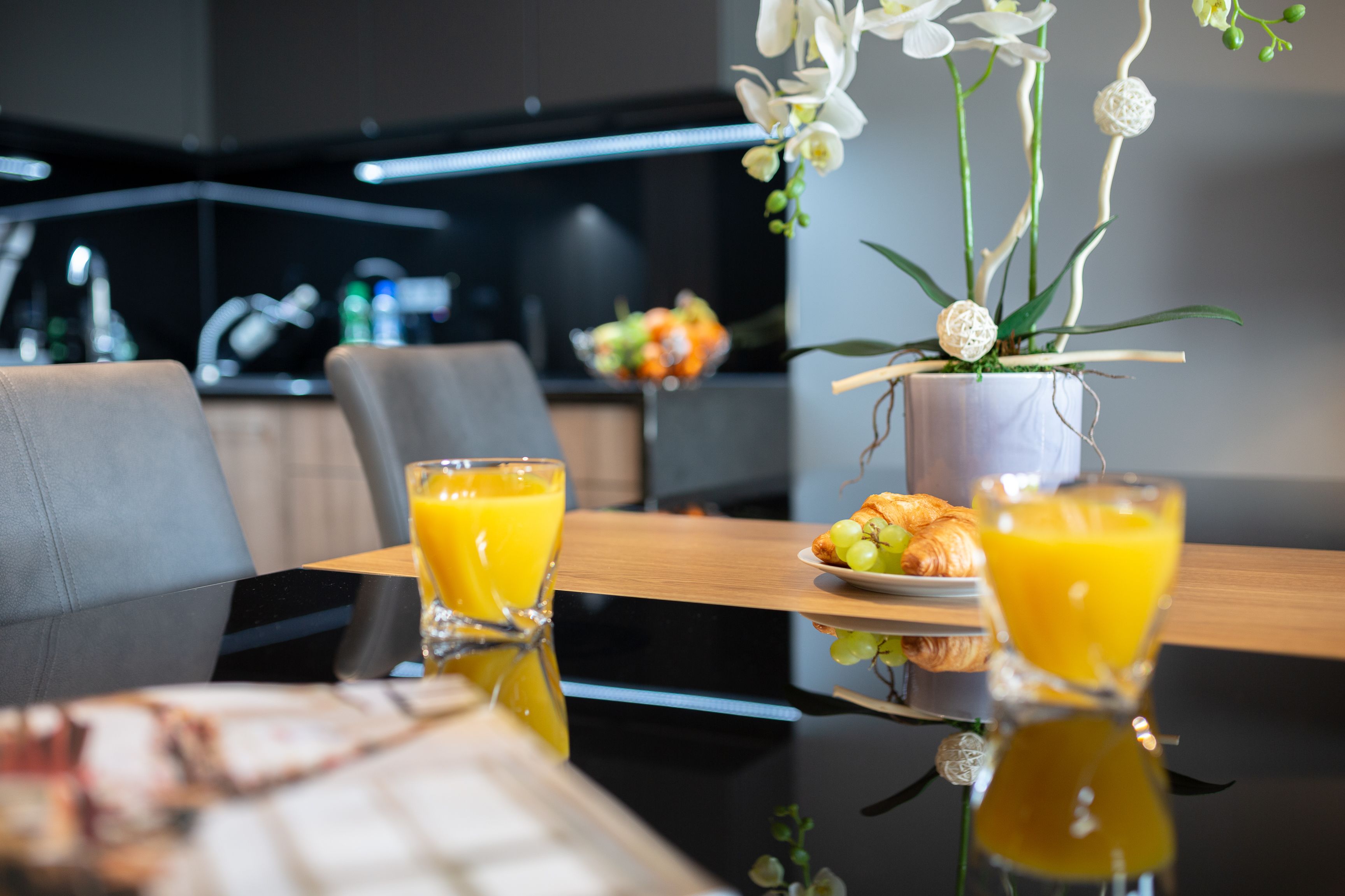 Una mesa de comedor con dos vasos de jugo de naranja, un plato con cruasanes y un florero con flores blancas.