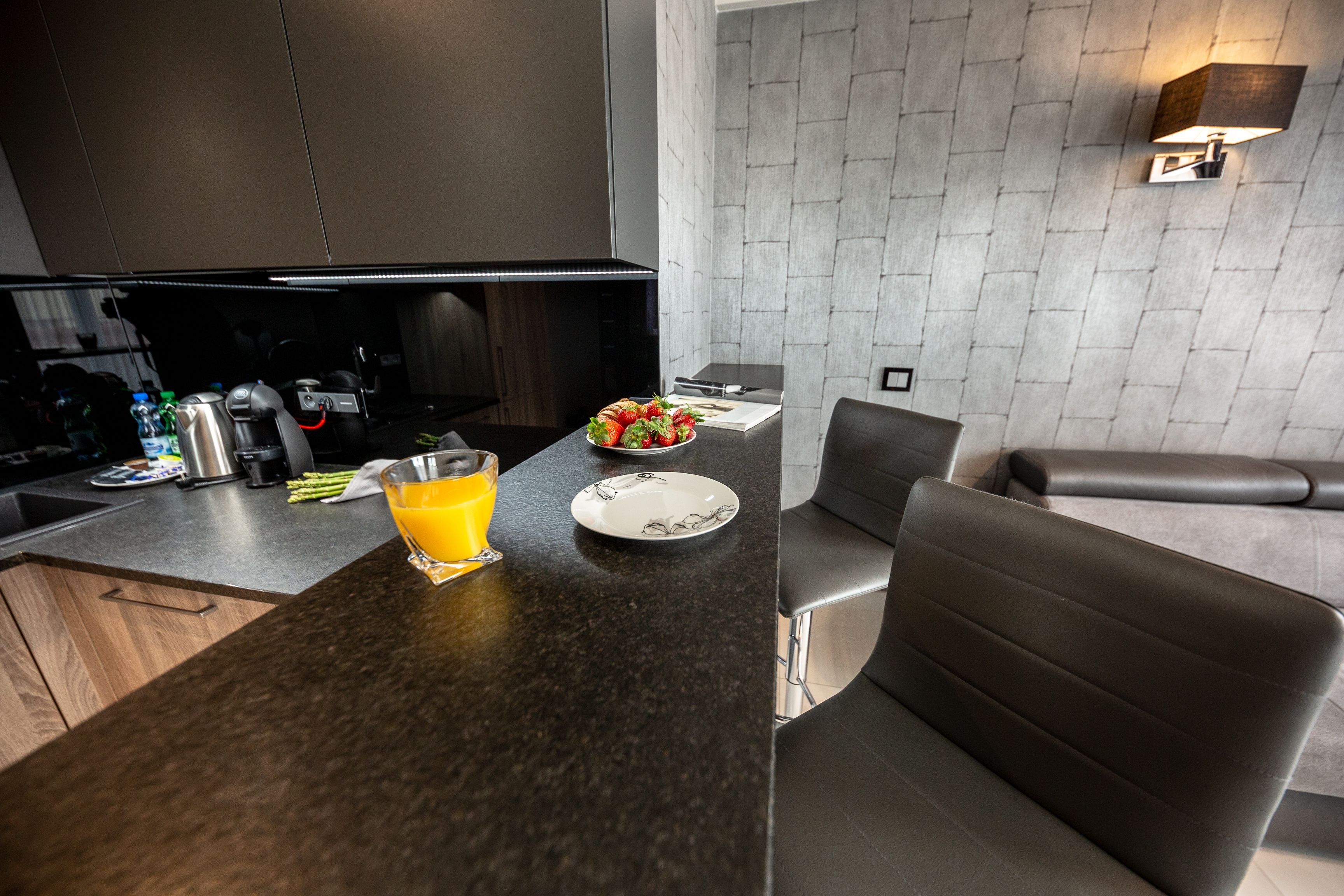A modern kitchen with a dark countertop, a glass of orange juice, a plate with food, and a dining area with a chair.