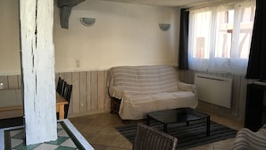 Una sala de estar con un sofá a rayas, una mesita y una silla. Se ve una ventana con cortinas y un radiador en la pared.