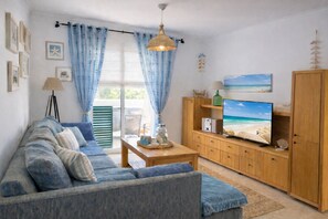 Ein Wohnzimmer mit einer Couch, Fernseher, Holzschrank und Blick auf den Strand durch das Fenster.