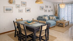 Een eethoek met een donker houten tafel en geweven stoelen, een blauwe bank met kussens, en een hanglamp van geweven materiaal.
