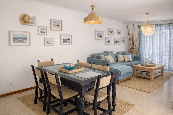 Ein Essbereich mit einem dunklen Holztisch und gewebten Stühlen, einer blauen Couch mit Kissen und einem hängenden, gewobenen Leuchter.