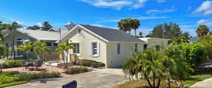 A residential house with a front porch, a small patio with outdoor furniture, and a well-maintained lawn with palm trees.