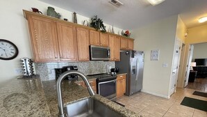 Cocina con encimeras de granito, pileta de acero inoxidable, muebles de madera, microondas, horno y refrigerador.