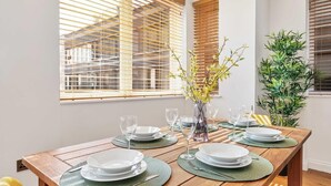A dining table set with plates, glasses, and a vase with flowers, in a room with wooden blinds and a plant. - Seaview Heights | By My Getaways (Brighton)