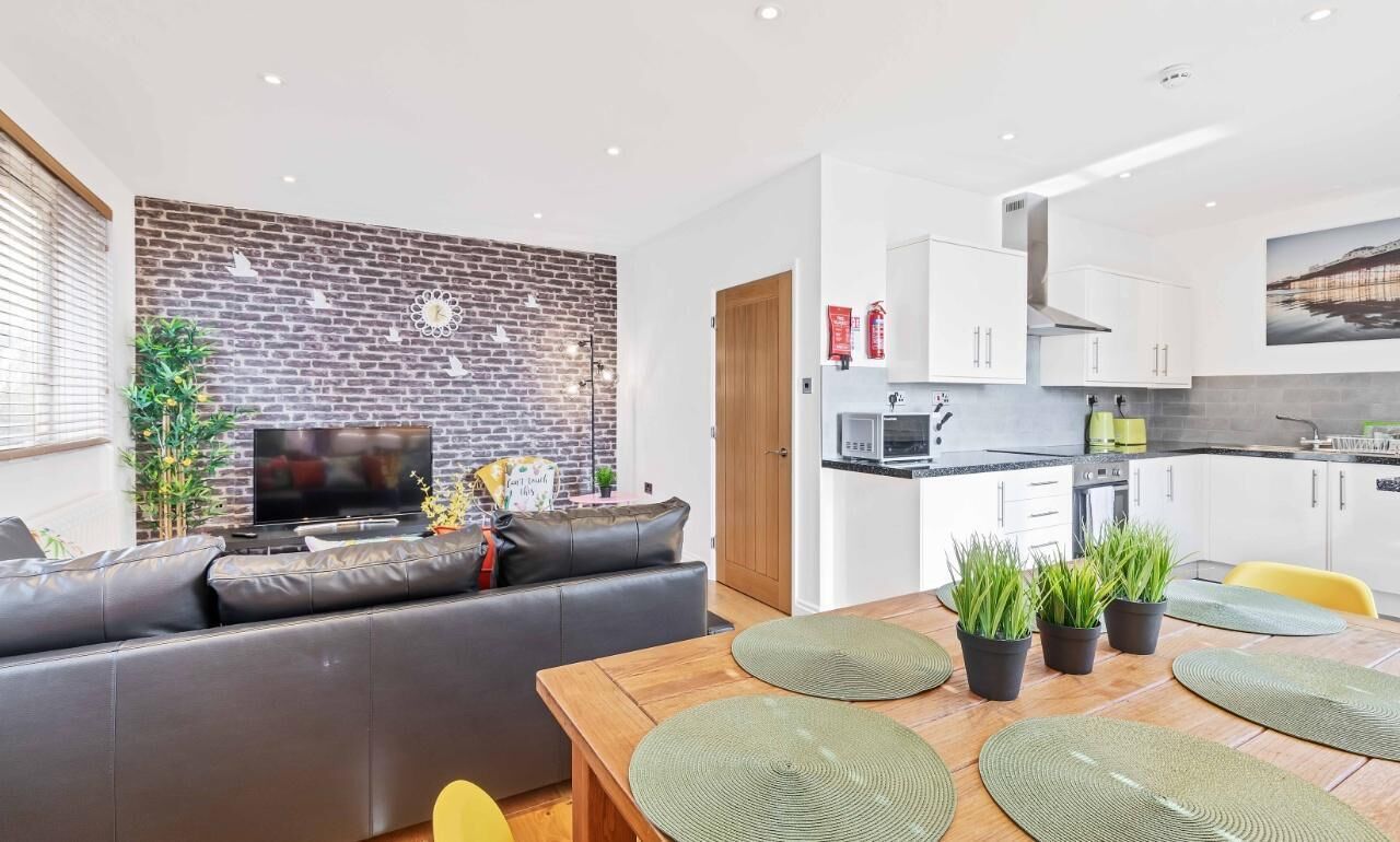 A modern living room with a brick wall, a flat-screen TV, a leather sofa, and a dining area with a wooden table and green placemats.