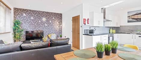 A modern living room with a brick wall, a flat-screen TV, a leather sofa, and a dining area with a wooden table and green placemats.