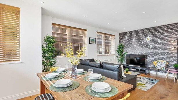 A modern living room with a black leather sofa, a dining table set for four, and a TV on a brick wall. - Seaview Heights | By My Getaways (Brighton)
