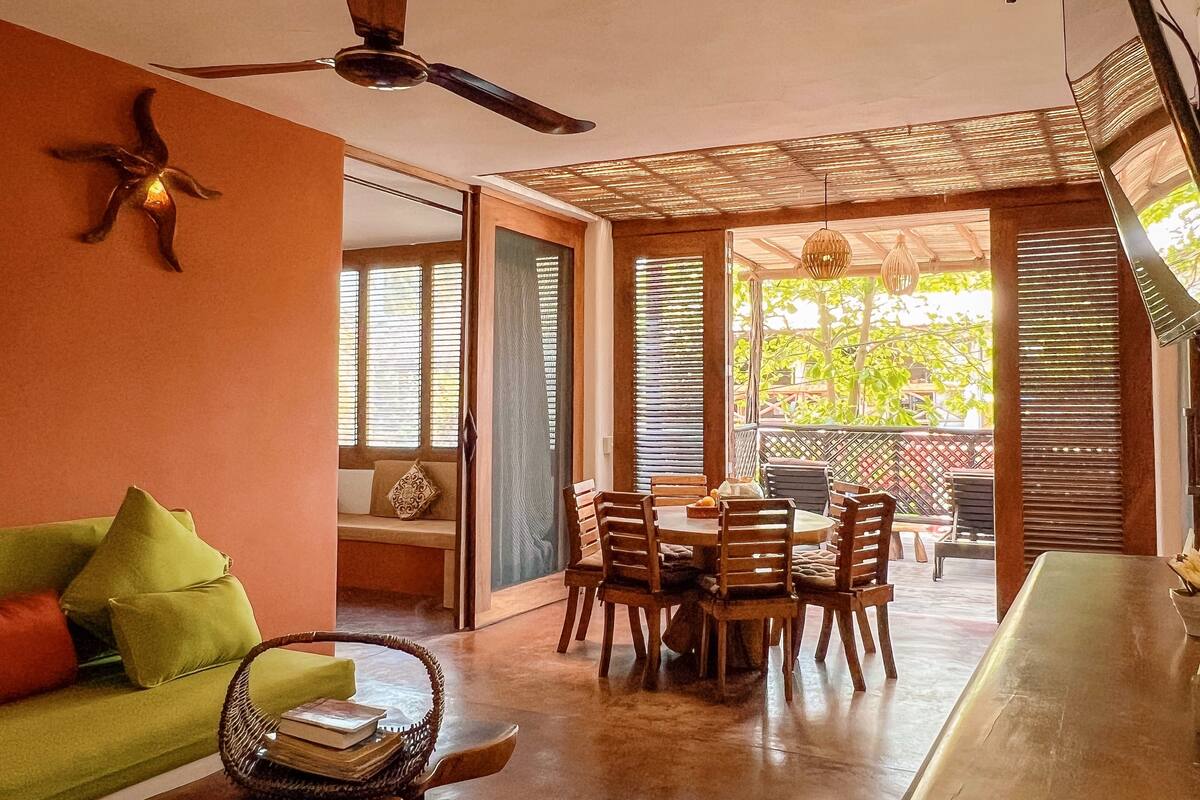 Una habitación con una mesa de comedor y sillas, un sofá verde, un ventilador de techo y vistas a través de una ventana con persianas.