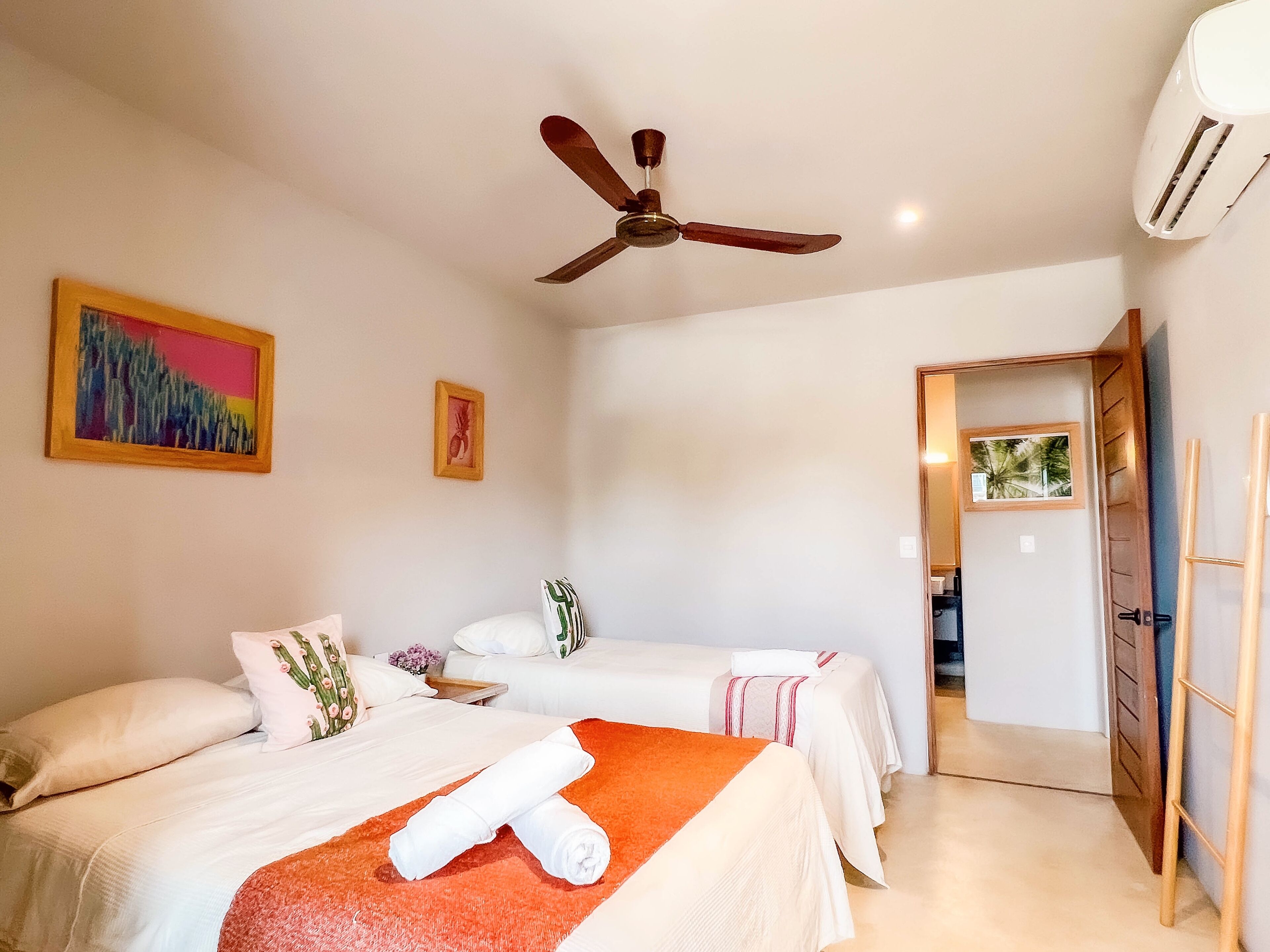 Habitación de hotel con dos camas, ventilador de techo, aire acondicionado y una puerta que da a otra habitación.