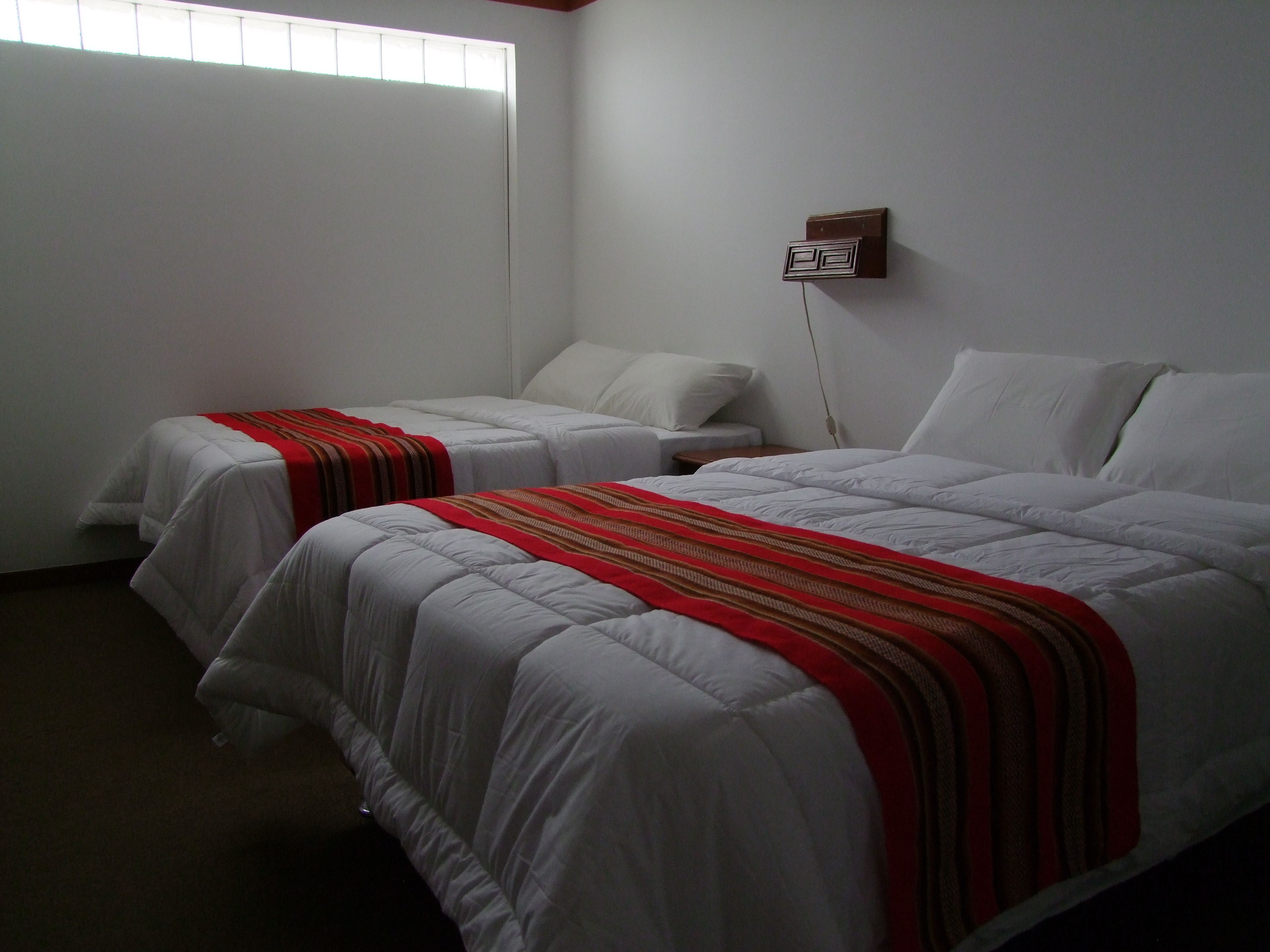 A hotel room with two single beds, a nightstand, a wall socket, and a window with blinds.