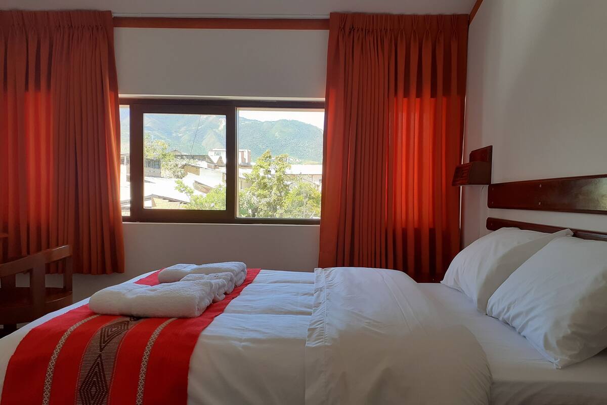 A hotel room with a bed, a window with curtains, and a view of the outside.