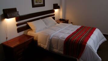 Una cama bien hecha con edredón blanco y manta a rayas marrón y roja, flanqueada por mesitas de noche de madera con lámparas, en una habitación con pared blanca lisa y un cuadro enmarcado sobre la cama.
