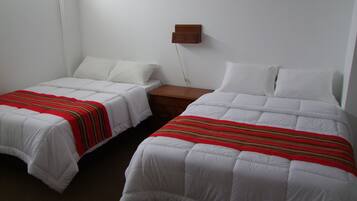 Dos camas individuales con ropa blanca y mantas a rayas rojas, un mesita de noche de madera y un enchufe de pared.