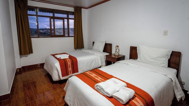 Habitación de hotel con dos camas, piso de madera, mesita de noche con lámpara y ventana con vista a un pueblo.