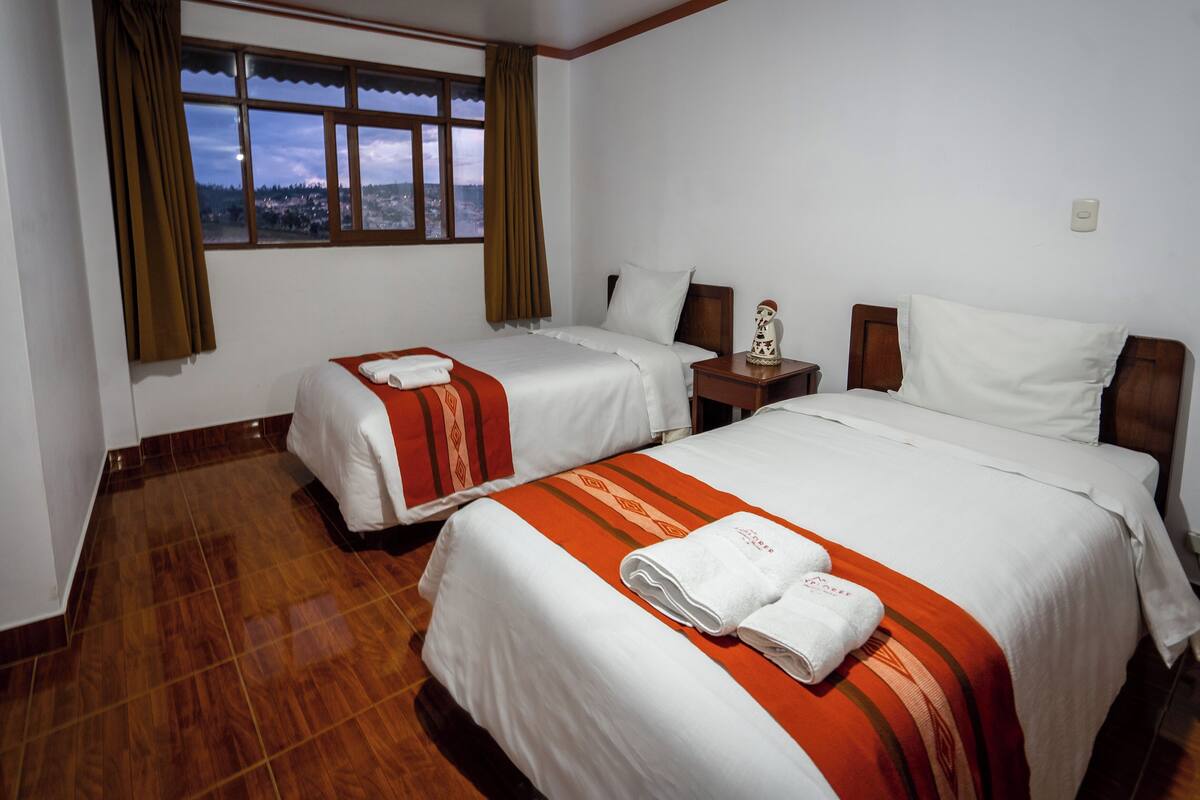 A hotel room with two beds, a wooden floor, a nightstand with a lamp, and a window with a view of a town.