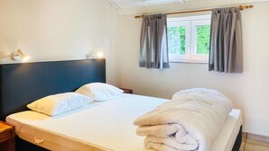 A neatly made bed with white linens and a rolled-up blanket, a wooden nightstand with a lamp, and a window with grey curtains.
