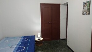 Een kleine, smalle kamer met een bed, een klein tafeltje met een lamp, een deur en een schilderij aan de muur.