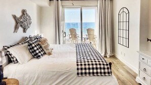 A bedroom with a large bed, checkered bedspread, a view of the sea through a sliding door, and a small balcony with chairs. - “Barefoot Bliss” - Two GulfFront Masters w/balcony (Panama City Beach)