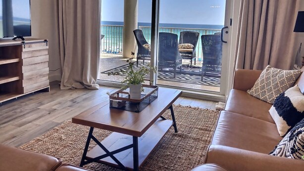 A living room with a sofa, a coffee table, and a TV. There is a balcony with chairs and a view of the ocean. - “Barefoot Bliss” - Two GulfFront Masters w/balcony (Panama City Beach)