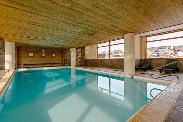 Une piscine intérieure dotée d’un plafond en bois et de murs en pierre, de grandes fenêtres et offrant une vue sur un paysage enneigé.
